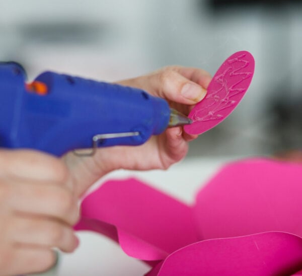 A blue glue gun spreading glue onto a hot pink paper flower petal.