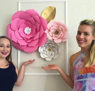 Two young women pose next to three giant paper flowers hanging on a white wall within a large white frame. The flowers are pink and silver with one gold accent leaf.