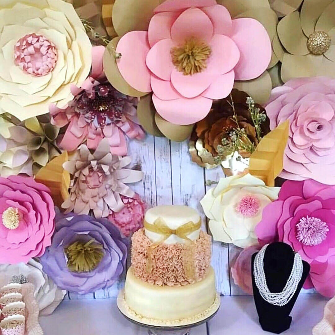 A pink and gold paper flower backdrop surrounds this three-tier birthday cake. The flowers are all different types in shade of pink, gold, cream, and purple.