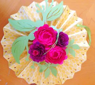 The image shows a yellow and pink paper fan rosette craft, with paper flowers and green paper leaves decorating the center. 