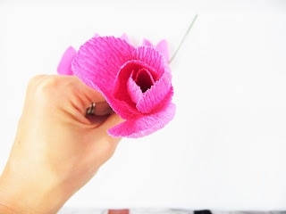 Abbi Kirsten holds a partially completed crepe paper rose in her hand, holding it close to the camera to show how the petals wrap around each other. Other petal pieces are in the background. 