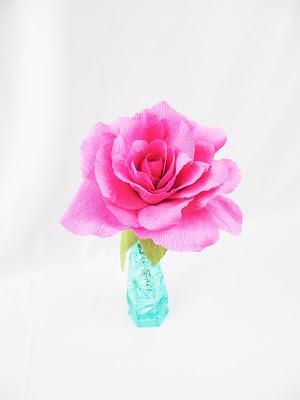 A blossoming pink crepe paper rose, with paper leaves, is in a small turquoise vase on a white background.