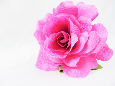 A closeup image of a pink crepe paper rose with small green leaves, resting on a white background. 