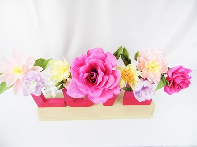 A bird's eye view of a crepe paper flowers in a variety of shapes and colors. Some of the letters that spell "love" are visible beneath the crown of flowers, which all sit on a white background cloth. 