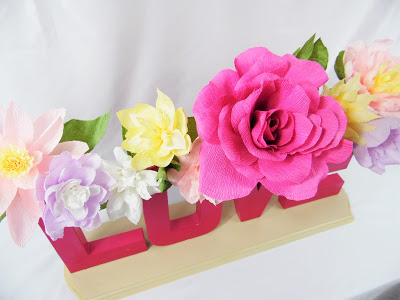 A close-up image of the colorful crepe paper flowers in shades of pink, lilac, yellow, and white that fill a cardboard typography vase. The letter vase spells the word “LOVE” and is attached to a wooden base that sits on a white table.