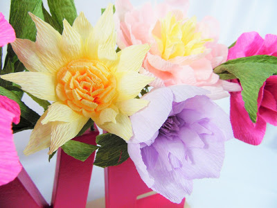 A bouquet of delicate, pastel-colored crepe paper flowers made using a Cricut cutting machine are seen on top of some wooden and cardboard letters. 