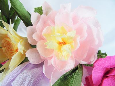 A closeup image of a light pink crepe paper flower with a yellow center. The flower is one of many colorful crepe paper flowers in a bouquet.