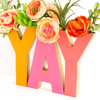 A DIY cardboard letter vase that spells out “YAY” in colorful letters. The tops of the letters are filled with stems of peony flowers, succulents, and green leaves.