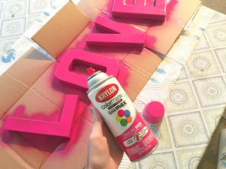 A person holds a can of pink Krylon spray paint. In the background, cardboard letters L, O, V, and E are painted pink, laying on a flat cardboard box.