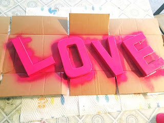 A closeup image of cardboard letters that spell out the word “LOVE,” spray painted pink and drying on a broken-down cardboard box. 