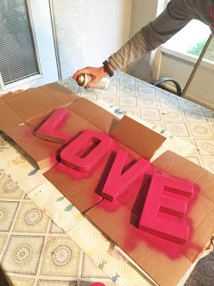 Abbi Kirsten demonstrates how to spray paint typography  letters for an even coat and vibrant color. The letters are on a table and cardboard, both used to protect the table from the paint. 