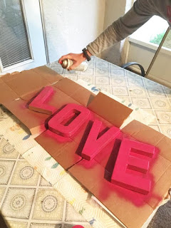 A person spray pains four cardboard letters with bright pink spray paint. The letters spell out the word LOVE.