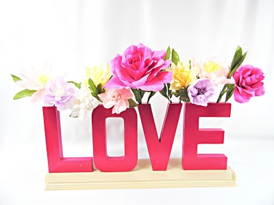 Red letters on a wooden base spell the word "Love." The top of each letter acts a a vase and holds crepe paper flowers. 