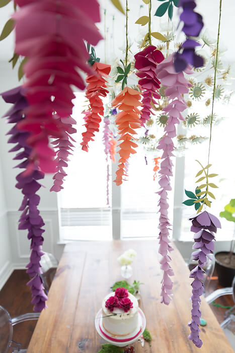 Colorful paper wisteria flowers hang from the ceiling over a table. A small single tier cake sits in the center of the table below.