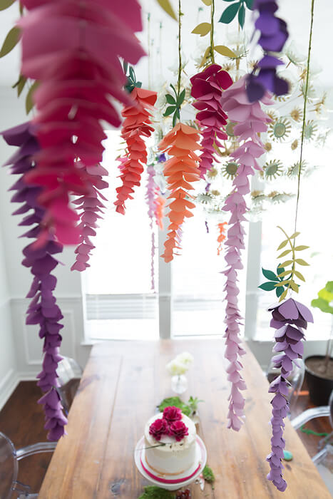 Colorful paper wisteria flowers hang from the ceiling over a table. A small single-tier cake sits in the center of the table below.