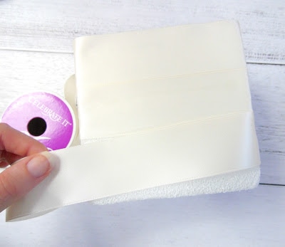 Abbi's fingers are seen carefully wrapping a floral block with wide satin ribbon while it rests on a white table. This is the base for the DIY wedding centerpiece craft. 