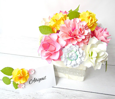 The finished DIY paper flower DIY wedding centerpiece is shown in an angle on top of a white wood table. The rhinestone ribbon faces outward and the paper flowers are in a beautiful arrangement above. A small yellow paper flower with paper greenery sits at the side of the centerpiece, holding the nametag to be used on the dinner table. The tag reads, "Abigail."