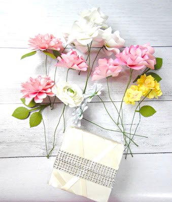 On a white wood table lays a cube wrapped in satin and rhinestone ribbons, with stemmed paper flowers above it. 