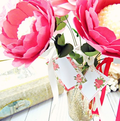 Stemmed paper peonies in a vase next to a white book. Mini Valentine's Day cards are attached to the paper flowers with satin ribbon. 