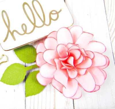 A pink paper Dahlia flower sits next to a small white trinket dish with the word "hello" painted in gold.