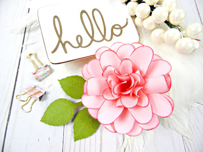 A pink ruffle-style paper dahlia flower sits on a wooden surface next to a white card that says "hello" in gold print and a string of pearls.