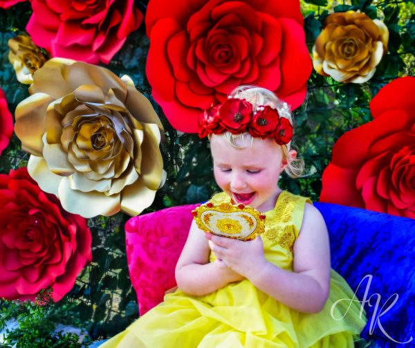 A little girl dressed as Belle looking into a gold mirror with large red and gold paper roses along green ivy as a backdrop. 