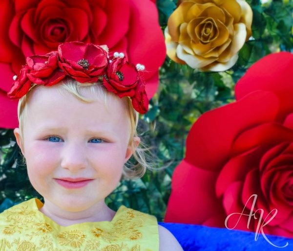 A little girl wearing a red flower crown with giant red and gold paper roses in the background. 