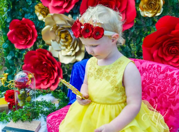 A little girl in a yellow dress wearing a red paper poppy crown looking into a golden mirror. Giant red and gold paper roses against green ivy are in the background. 