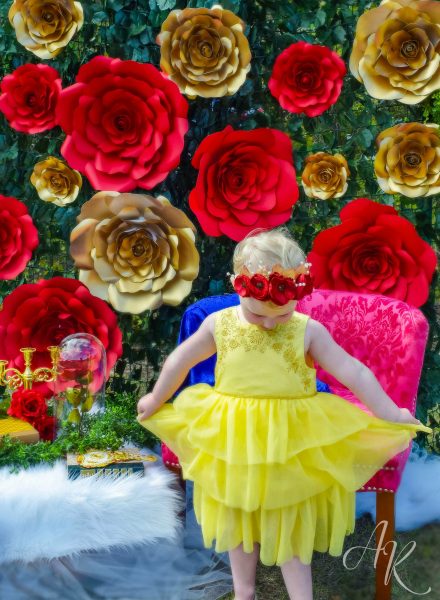 A child in a yellow dress and red poppy crown holding out her dress. In the background there is a wall of green ivy covered with giant red and gold paper roses. 