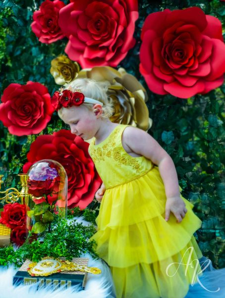 A little girl in a yellow dress looking into a glass dome containing a single red rose. In the background, there are giant red and gold paper roses along green ivy. 