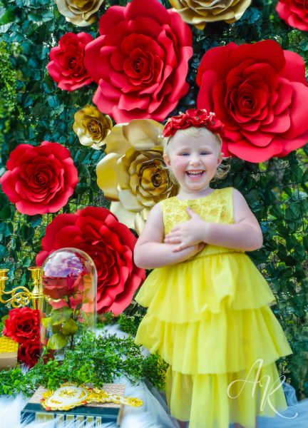 A smiling girl in a yellow dress and red paper poppy crown. The background is filled with giant red and gold paper roses along green ivy. 