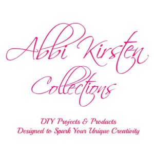 Pink script font on a white background says "Abbi Kirsten Collections". A smaller line of pink text says "DIY Projects & Products Designed to Spark Your Unique Creativity"