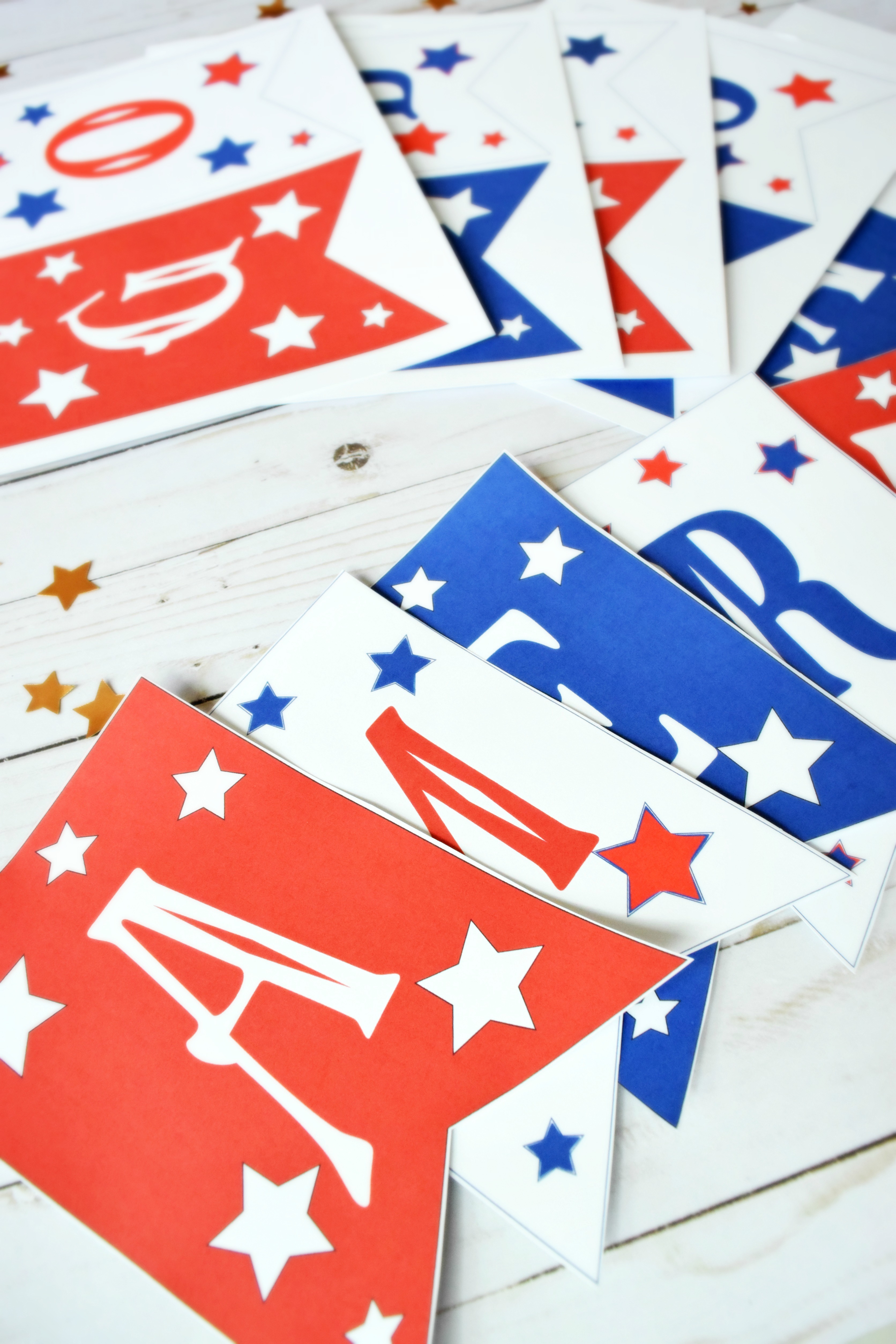 Red, white, and blue paper banner flags are laid out on a white wood table, each with a letter and stars on the front. This banner spells out "god bless america".