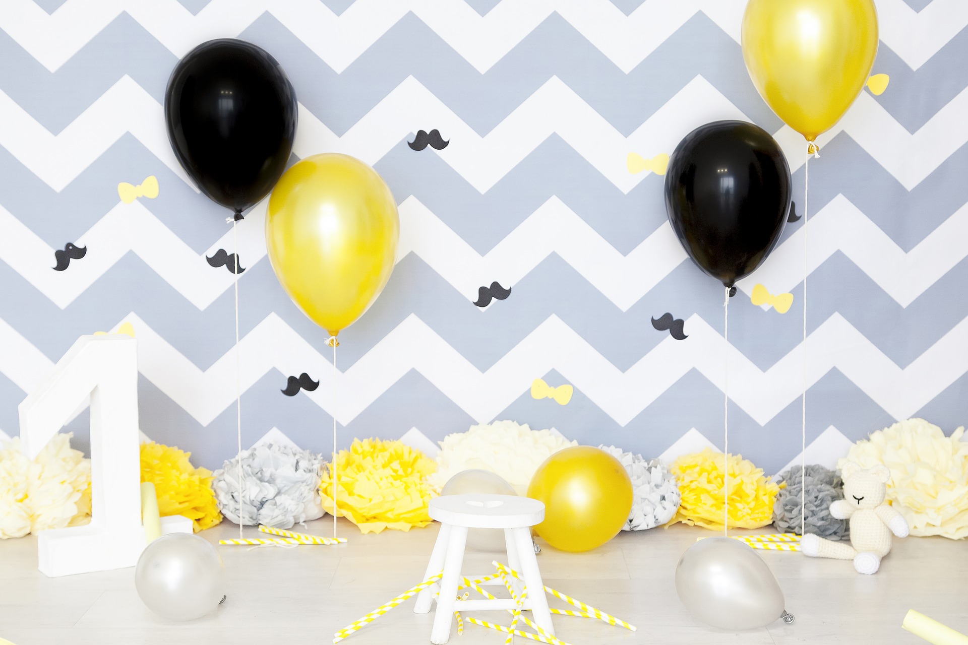 A photo background for a 1 year old's birthday party. Gold and black balloons float in the air, and yellow, gray, and white paper flowers sit on the floor in front of a decorative blue and white zig-zag striped wall.