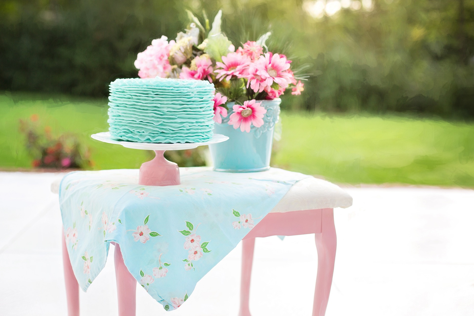 A single tier birthday cake with teal blue icing sits on a cake platter, posed on a pink and white bench with a bucket full of pink flowers.