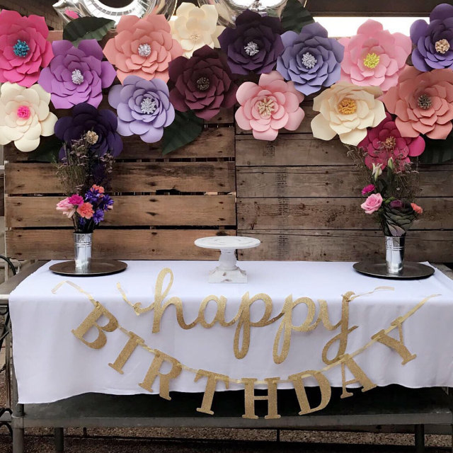 A table covered in a white tablecloth is decorated with a gold "happy birthday" banner and flower arrangements. On the wooden wall behind a table, a giant paper flower banner hangs, made up of colorful paper flowers.