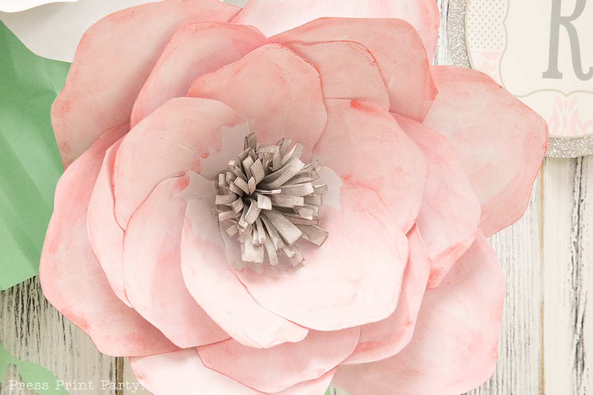 A closeup image shows a large pink paper flower with a fluffy flower center, blooming petals, some paper leaves, and a white wooden background. These paper flowers are designed by Abbi Kirsten and made by Nathalie Brown from Press Print Party for a shabby-chic baby shower. 