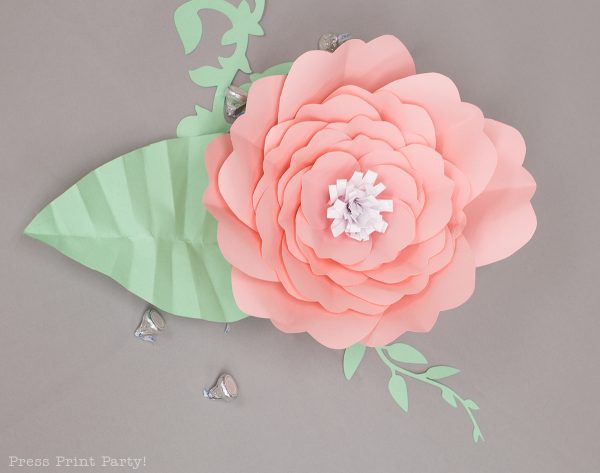 An overhead view of a pink paper flower with a white center and paper greenery on a gray background. The petals have wavy edges, which adds softness to the paper flower. 