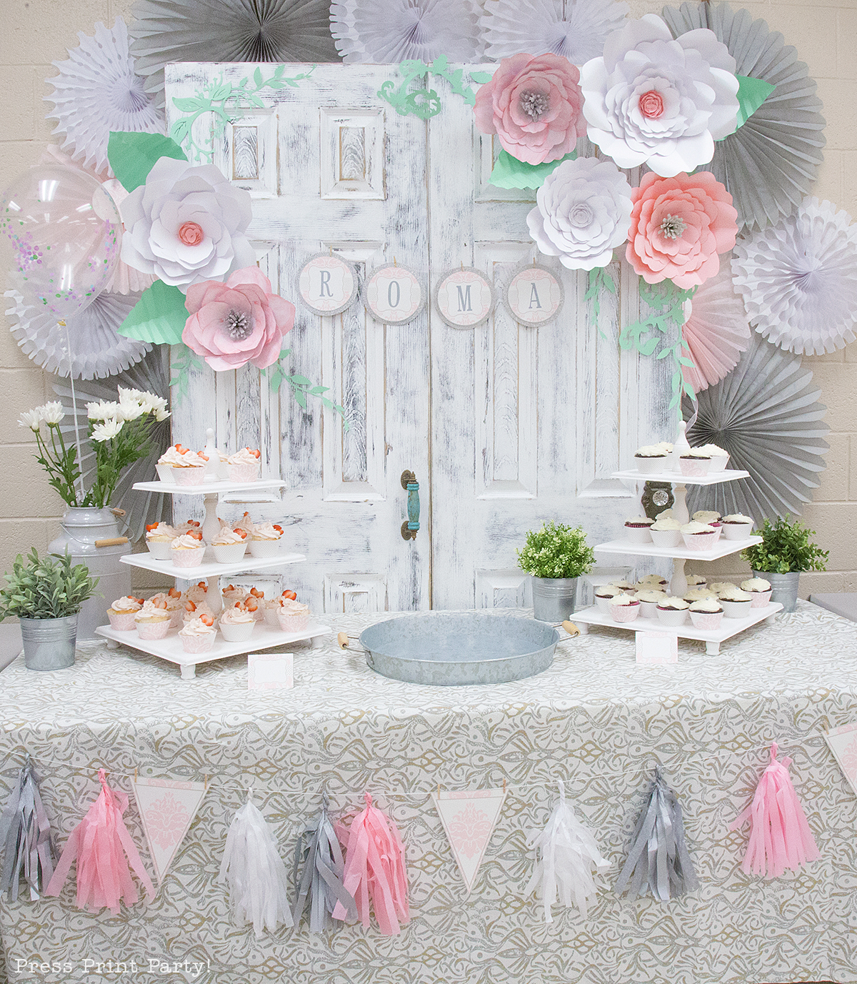 A table decorated for a baby shower. There are large paper flowers, a banner made from tassels and triangle flags, a white wooden backdrop on which another banner is hung, and decorative paper fans adorn the walls. These designs can be found on Abbi Kirsten’s blog. 