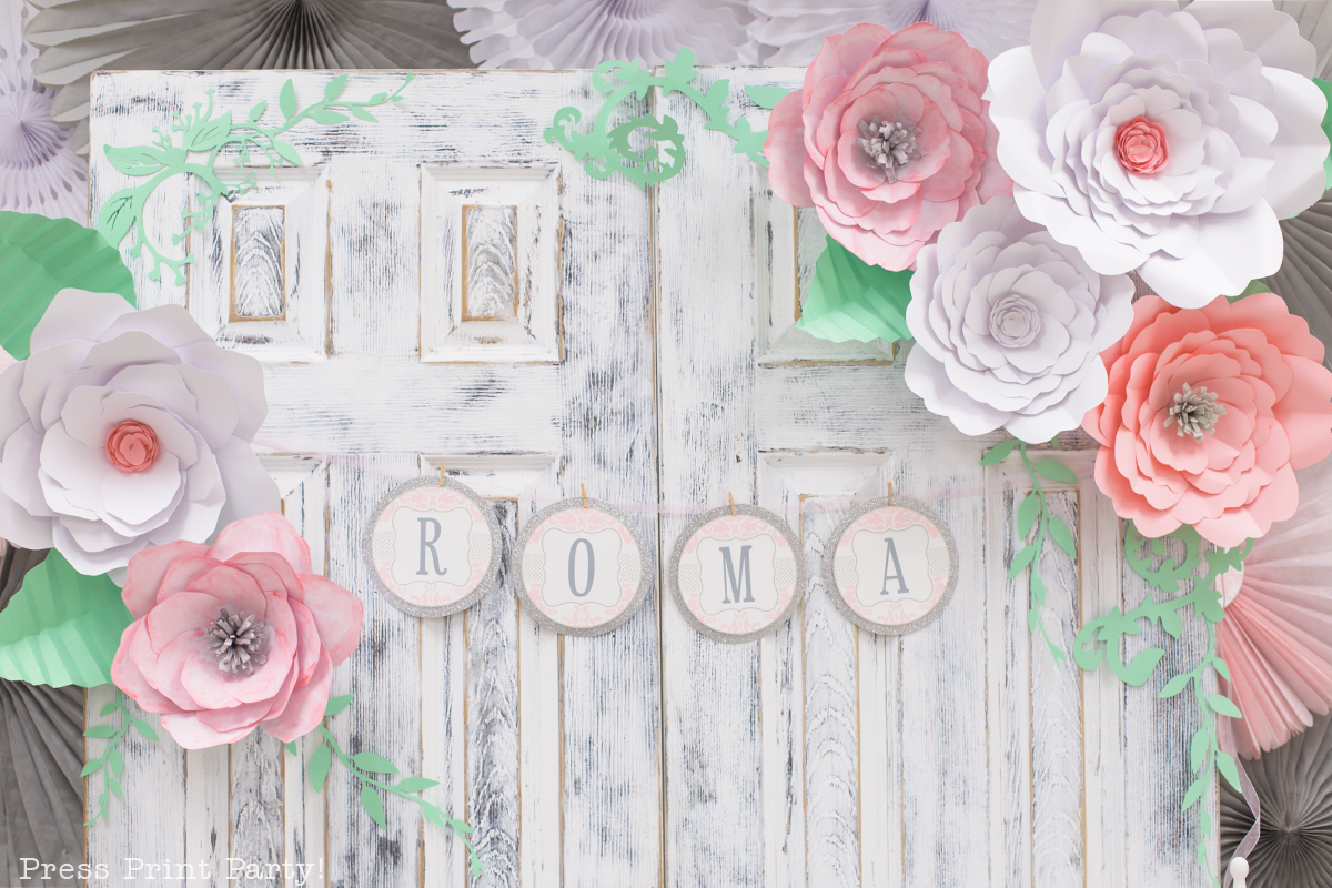 A closeup of two rustic wood used as a backdrop for a dessert table at a baby shower. The wood is white, distressed, and decorated with a banner that reads "Roma" and large, delicate paper flowers. 