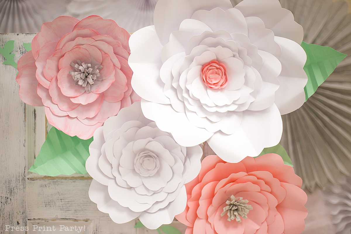 An overhead picture of four paper flowers in large and giant sizes, with paper greenery on a wooden background. Each paper flower was created using templates designed by Abbi Kirsten and made the perfect flowers for baby shower decorations. 