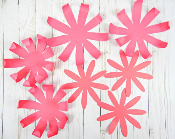 Seven pink paper Chrysanthemum  flower petals sit on a whitewashed wood work surface. There are four larger petals, and three medium sized petals. 