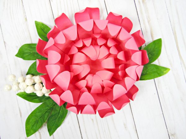 A large pink Chrysanthemum paper flower with green leaves sits on a white-washed wood work surface. Learn how to make these gorgeous pink paper flowers with this easy tutorial.