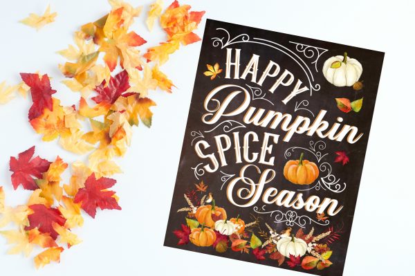 The dark colored "Happy Pumpkin Spice Season" free fall printable lays at an angle on an off-white background next to beautiful fall-colored leaves.  