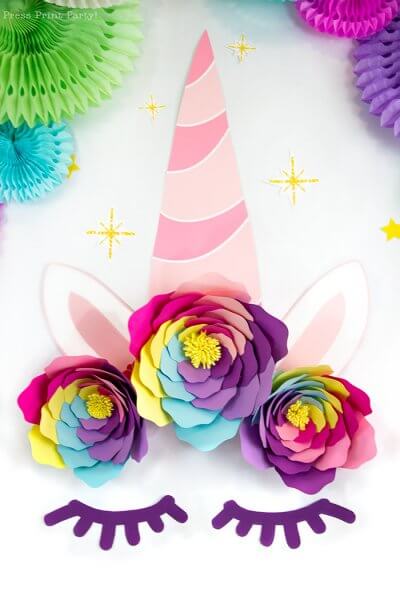 A paper unicorn backdrop with thick lashes, a pink horn and three beautiful iris rainbow flowers that make a crown for the unicorns head. 