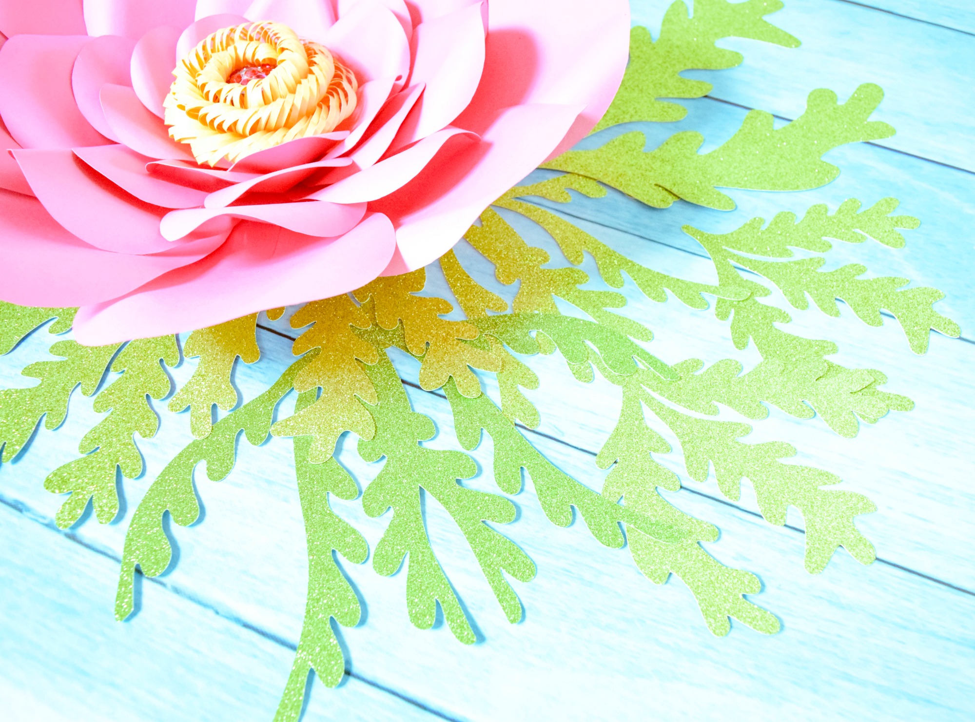 Branches of green glittery seaweed flow out from under a large pink paper flower, spread out on a blue table.