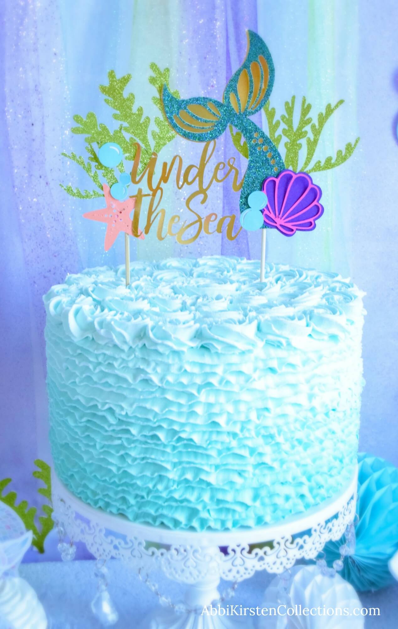 A light blue cake is frosted to look like ocean waves and is displayed in front of a blue and green backdrop reminiscent of the ocean. The large topper says "Under the Sea" in gold and has a starfish, shell, bubbles, and a mermaid tail.