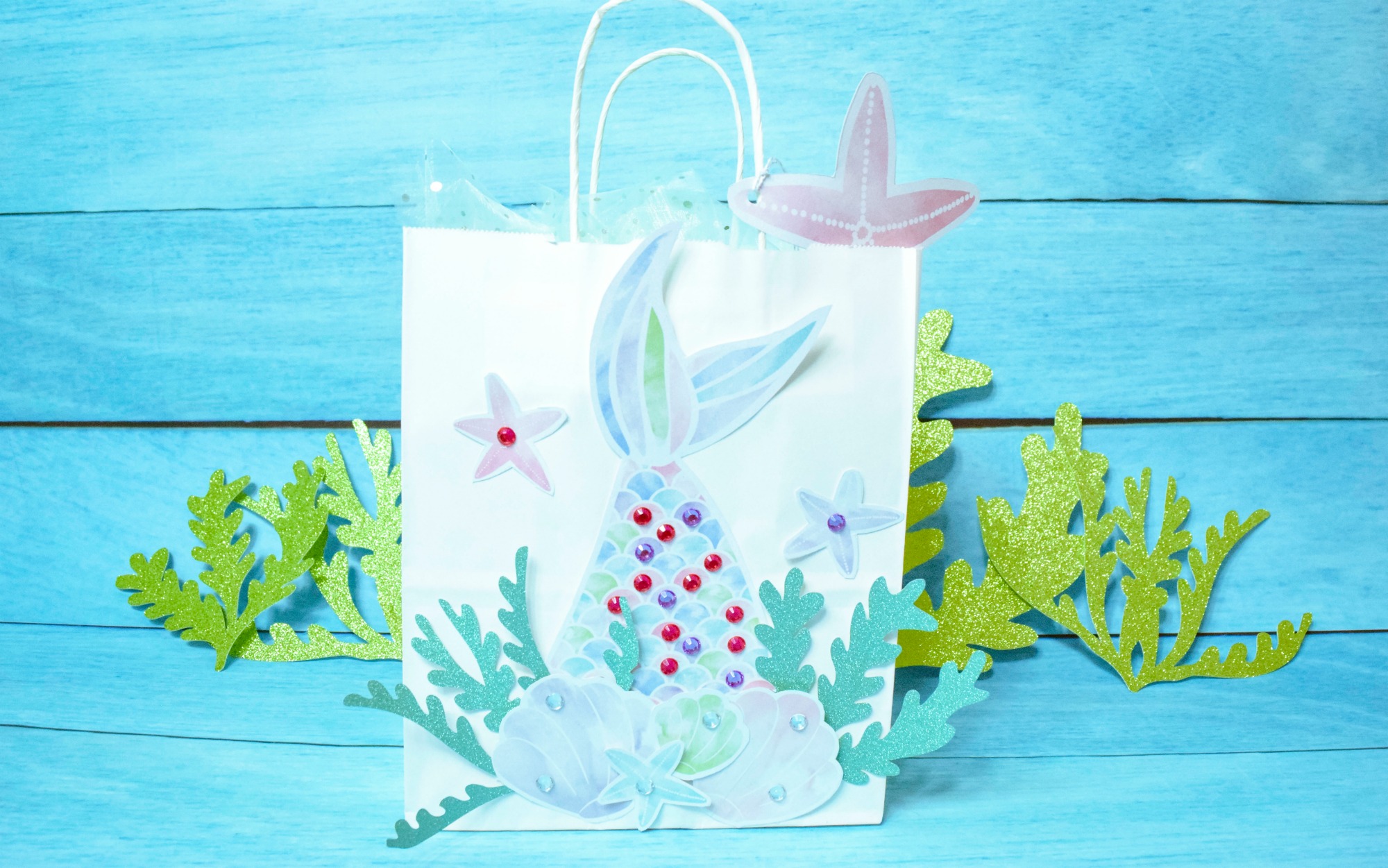 A DIY mermaid gift bag created with colorful paper cutouts - a mermaid tail, seaweed cutouts, seashells, and starfish.