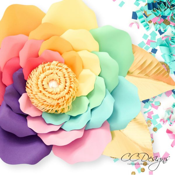 A closeup of a beautiful large rainbow paper flower with various colors of petals swirling to the golden center of paper fringe. Yellow-gold leave peek out from under the flower. The papercraft sits on a white background with colorful party confetti. 
