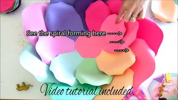 An ad for the DIY Iris Unicorn Rainbow Flowers and Large Rainbow Paper Flowers video tutorial. The text also includes arrows pointing to the paper flower petals with "see the spiral forming here."